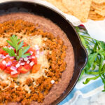 In a cast iron skillet there is a layer of pureed black beans, then cooked chorizo, melted cheese, tomatoes and chopped red onion.