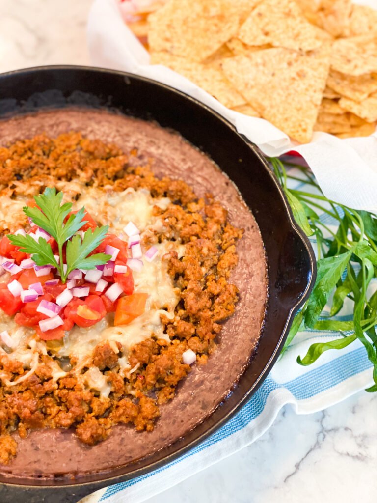 In a cast iron skillet there is a layer of pureed black beans, then cooked chorizo, melted cheese, tomatoes and chopped red onion.