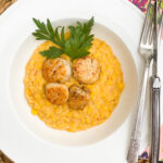 Creamed corn in the bottom of a round bowl with 4 seared scallops on top in the middle with a green parsley garnish.