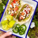 Two CHICKEN TACOS loaded with guac, tomatoes and cheese in a square serving dish and a garnish of limes and jalapenos.