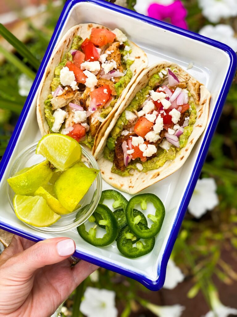 Two CHICKEN TACOS loaded with guac, tomatoes and cheese in a square serving dish and a garnish of limes and jalapenos.