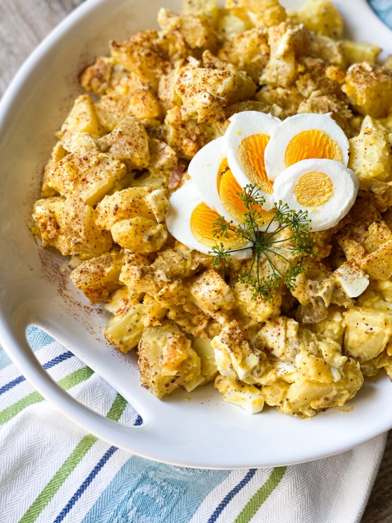 Potato Salad in a round white bowl with sliced egg and dill on top as a garnish.