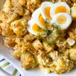 Potato Salad in a round white bowl with sliced egg and dill on top as a garnish.