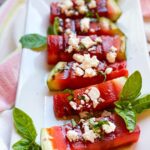 GRILLED-WATERMELON spears on a white rectangle platter with feta and balsamic vinegar on top and garnished with fresh basil.