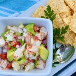 SHRIMP-CEVICHE in a square bowl with silver spoon and a side of tortilla chips.