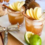 Two glasses of APPLE PIE MARGARITA. Light brown liquid the color of cinnamon is garnished with apples and cinnamon stick and a cinnamon sugar rim