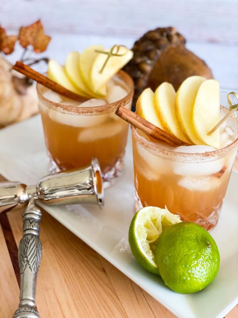 Two glasses of APPLE PIE MARGARITA. Light brown liquid the color of cinnamon is garnished with apples and cinnamon stick and a cinnamon sugar rim