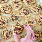 A cookie sheet full of chocolate chip cookies covered in pecans, melted chocolate and caramel drizzle