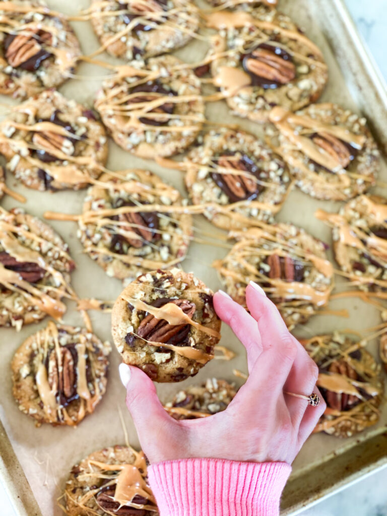 A cookie sheet full of chocolate chip cookies covered in pecans, melted chocolate and caramel drizzle
