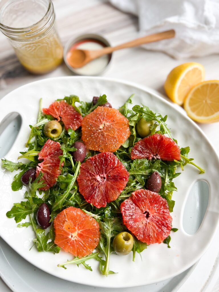 Bright colorful arugula salad topped with olives and two types of oranges.
